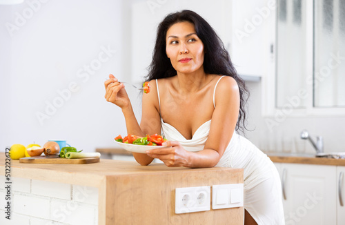 Portrait of asian woman in nightie eating salad in kitchen at home. Woman leaning on kitchen table, eating salad. photo