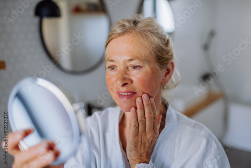 Beautiful senior woman in bathrobe looking at mirror and applying natural face cream in bathroom, skin care concept.