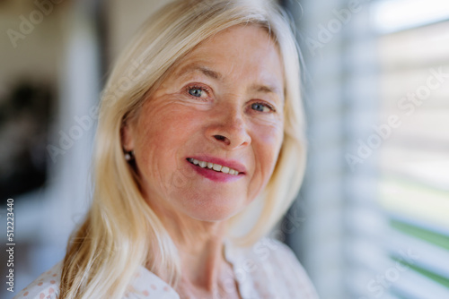 Portrait of attractive senior woman standing by window at home, looking at camera and smiling.