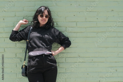 happy young black-haired woman dressed in black sweatshirt and black pants and sunglasses leaning against a mint colored brick wall grabbing her hair and smiling