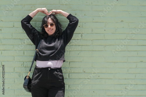 happy young black haired woman dressed in black sweatshirt, black pants and sunglasses leaning against a mint colored brick wall making a heart with her arms and smiling