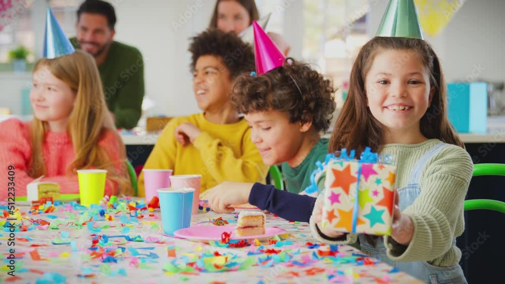 Girl sitting at table for party celebrating birthday holding gift out towards camera - shot in slow motion Girl sitting at table for party celebrating birthday holding gift out towards camera - shot in slow motion