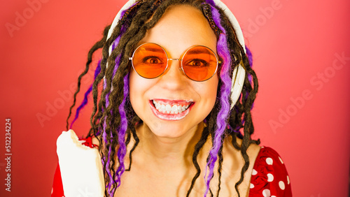 Young woman in sunglasses, headphones standing with bitten ice cream, smiling, looking at camera. Cheerful female with dreadlocks, her lips in sundae, listening to music in studio.