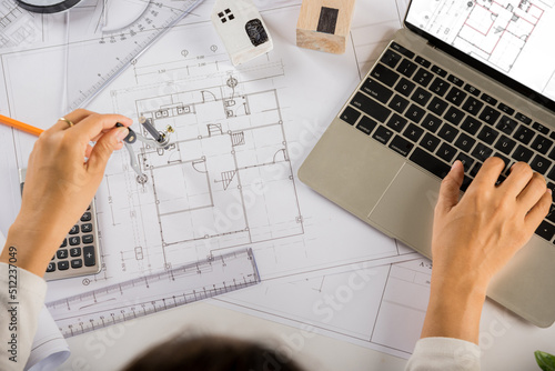 Architectural project workplace. Architect typing laptop keyboard to review design of house building before editing or sketching with compass on blueprint paper on table desk at architecture office