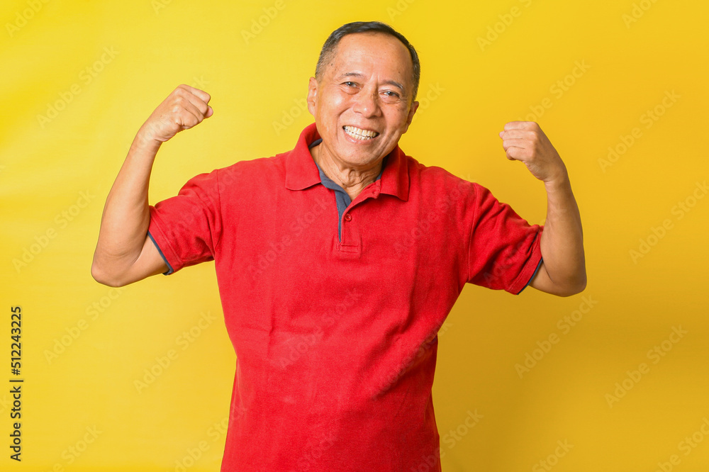 Asian pensioner grandpa raise two hands show muscles wear red t-shirt isolated yellow color background Asian pensioner grandpa raise two hands show muscles wear red t-shirt isolated yellow color background