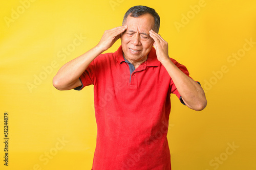 Asian senior man wearing casual t-shirt over yellow background suffering from headache and stressed because pain and migraine. Hands on forehead.
