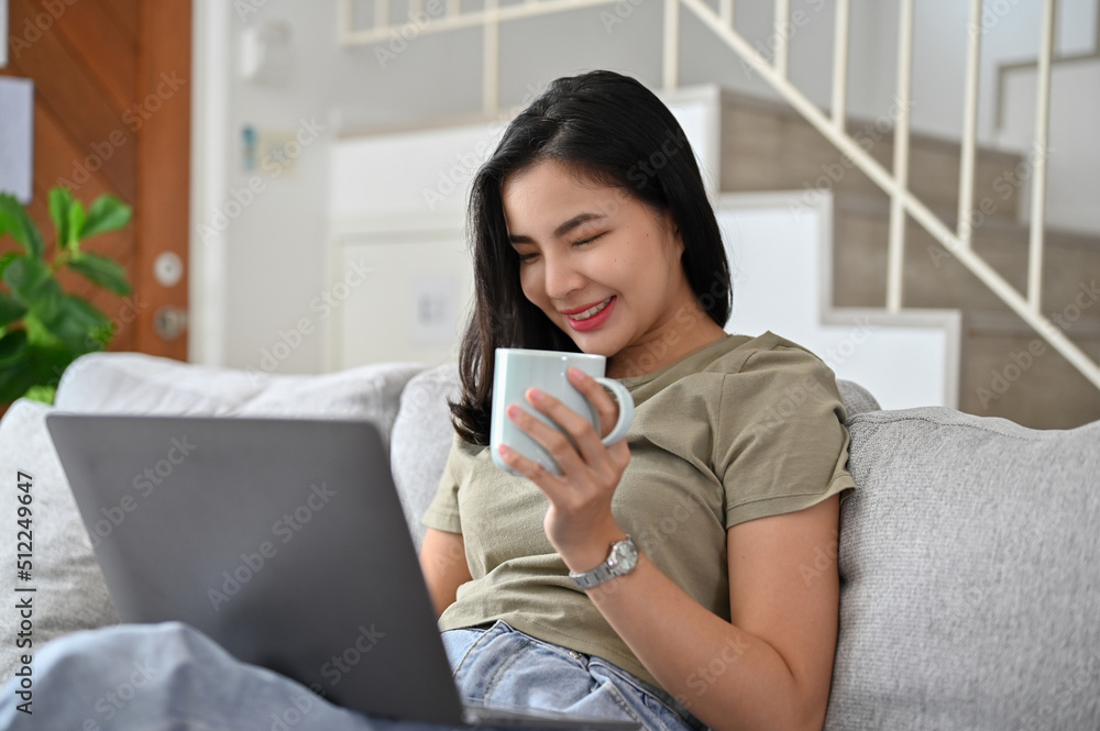 Beautiful young Asian woman having a coffee on the sofa and using laptop computer. Beautiful young Asian woman having a coffee on the sofa and using laptop computer.