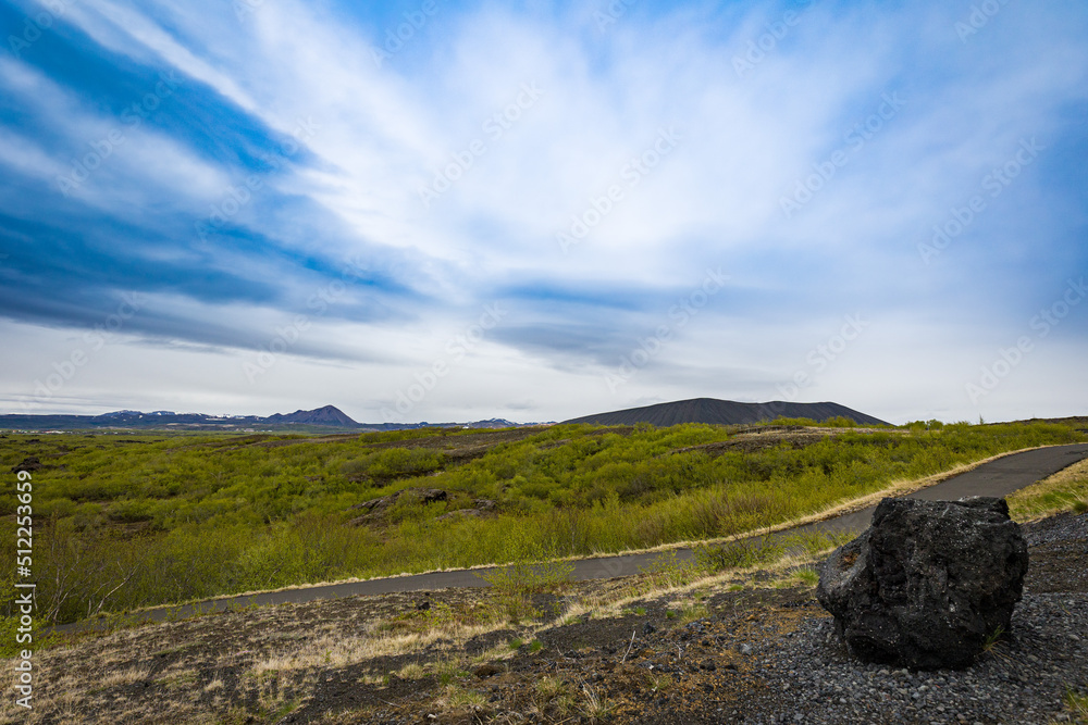 A chunk of volcanic rock in front of a pathway, a meadow and a crater in Northern Iceland