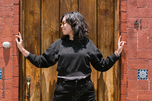photo of a cheerful young woman dressed in black posing in a fun way in front of a wooden door set in a wall with Mexican-style decoration