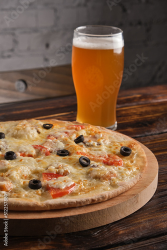 Cheese pizza with tomatoes and olives on a wooden platter and a glass of light beer on the table in the pub. Vertical orientation, close-up, copy space, no people