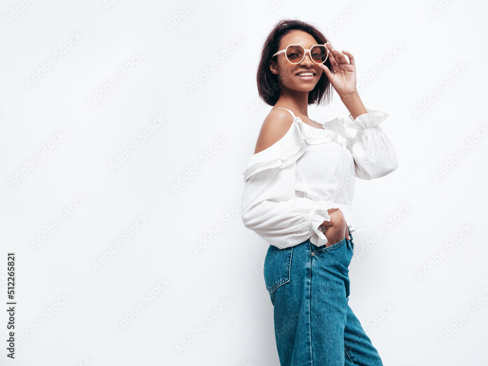 Portrait of young beautiful black woman. Smiling model dressed in summer jeans clothes. Sexy carefree female posing near white wall in studio. Tanned and cheerful. In sunglasses Portrait of young beautiful black woman. Smiling model dressed in summer jeans clothes. Sexy carefree female posing near white wall in studio. Tanned and cheerful. In sunglasses