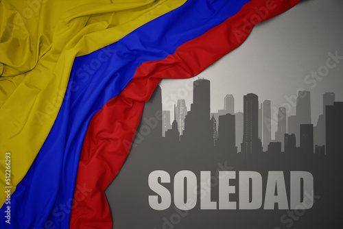 abstract silhouette of the city with text Soledad near waving national flag of colombia on a gray background.