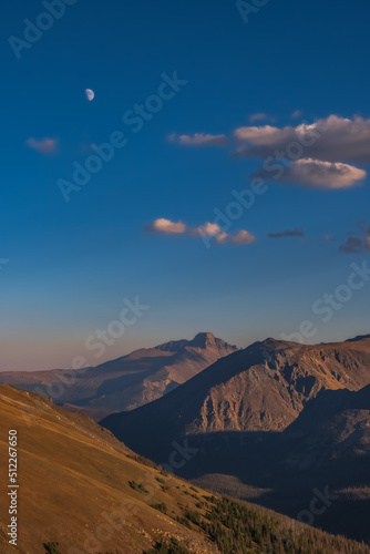 Vertical view of bare mountain peaks about tree line as seen from trail ridge road in Rocky Mountain National Park, Colorado, at sunset; blue sky with beautiful clouds and rising moon in background