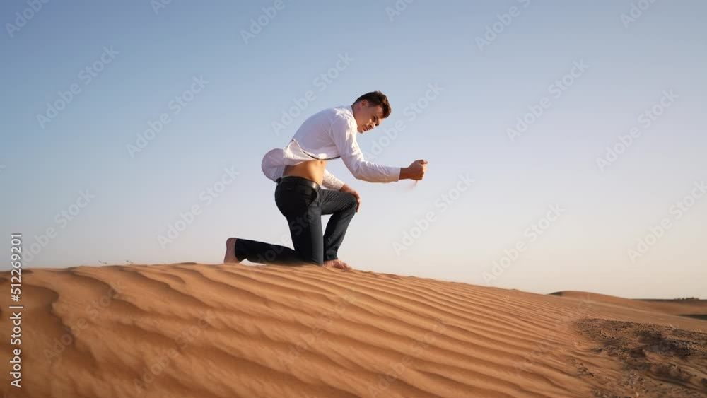 Male adult in white shirt and black pants taking a handful of sand in the desert at sunset and pouring it through the fingers Male adult in white shirt and black pants taking a handful of sand in the desert at sunset and pouring it through the fingers