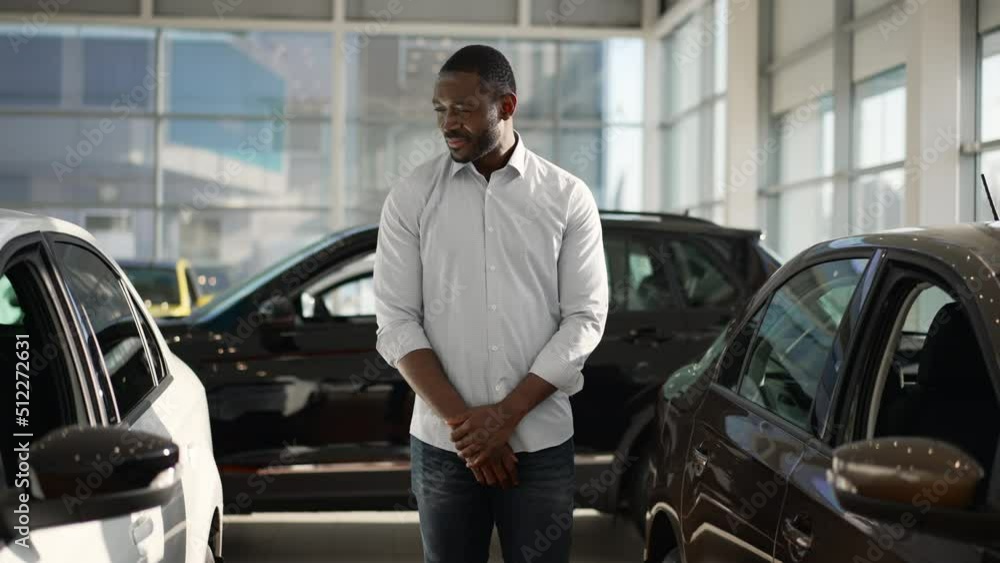 Young businessman is looking at new cars and posing for camera while standing in dealership spbas. 4k Portrait of handsome american african man studies transport models for sale and looks with smile Young businessman is looking at new cars and posing for camera while standing in dealership spbas. 4k Portrait of handsome american african man studies transport models for sale and looks with smile