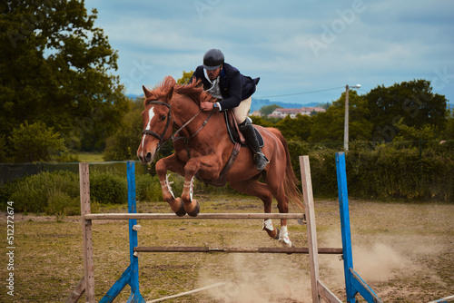 Dark-skinned horseman equipped with racing suit and helmet jumping an obstacle riding his brown horse