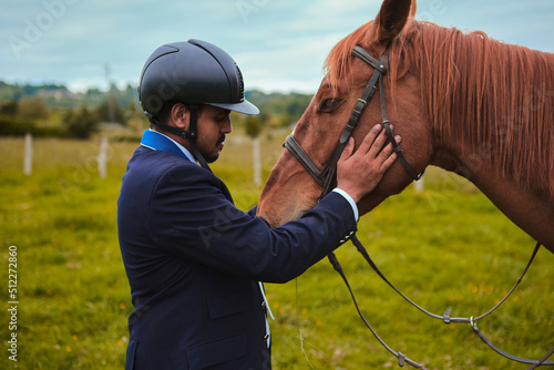 Ebony jockey caressing and loving his equine wearing helmet and competition suit on a green meadow
