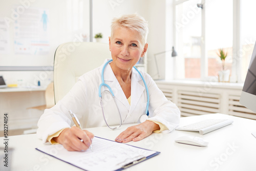 Portrait of smiling mature doctor with stethoscope on neck sitting at table and making notes in medical record