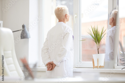 Pensive female director of Medicine wearing white coat holding hands behind back and looking out window while thinking of solution