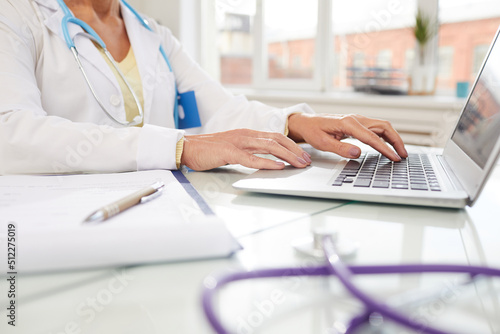 Close-up of unrecognizable female doctor in lab coat sitting at desk and examining information on laptop