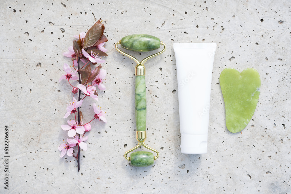 Obraz na płótnie Facial massage kit on stone concrete background - jade gua sha tool and face roller with cosmetic moisturizer or face cream tube and blooming sakura branch. Natural stone face massager, mockup w salonie