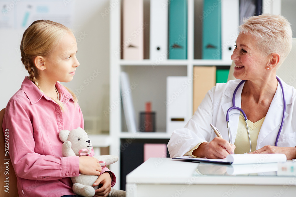 Obraz premium Cheerful attractive female pediatrician with short hair sitting at table and writing down in clipboard while talking to little patient