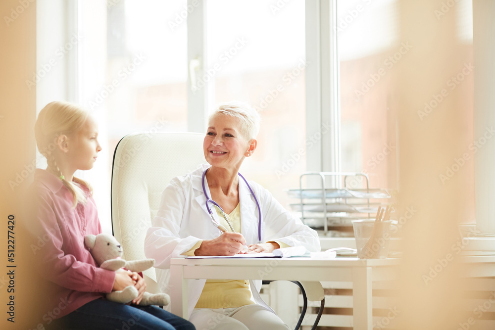 Content attractive female pediatrician in lab coat sitting at desk and making notes in medical card while asking child about complaints Content attractive female pediatrician in lab coat sitting at desk and making notes in medical card while asking child about complaints