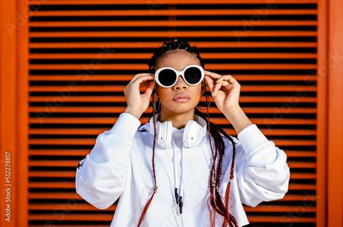 Young African American woman with braided hair wearing urban sportswear with headphones and sunglasses on an orange background