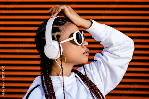 Young African American woman with braided hair wearing urban sportswear with headphones and sunglasses on an orange background photo