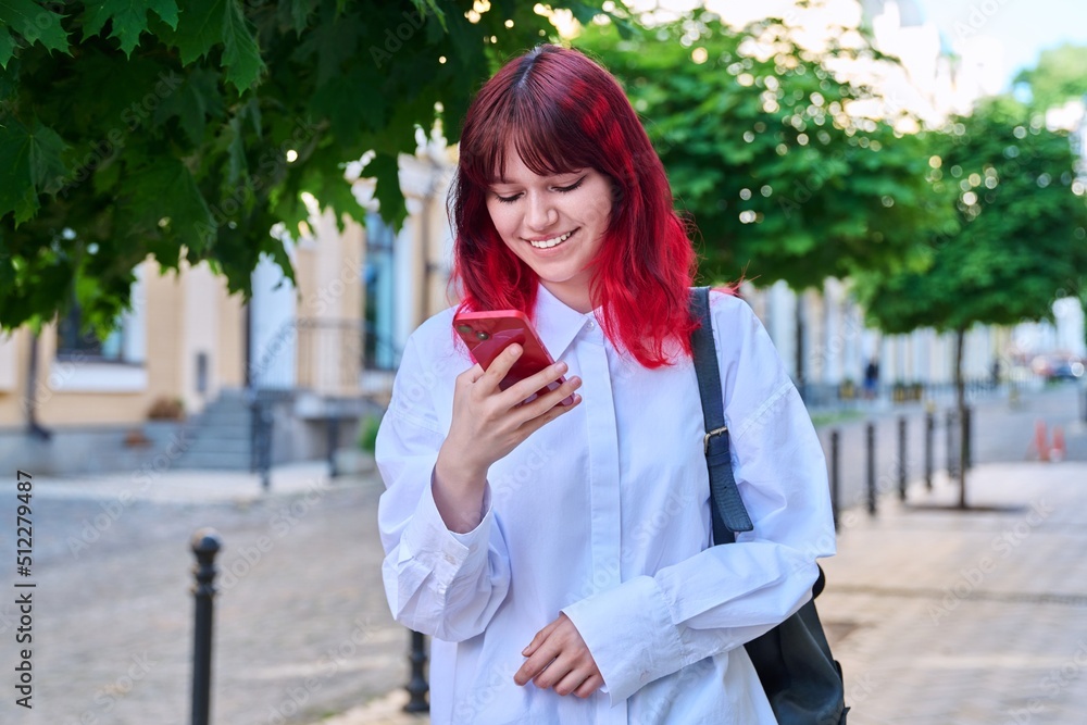 Fototapeta premium Beautiful smiling teenage female looking at phone on city street