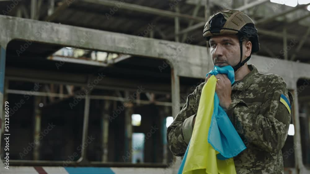Tired emotional soldier in camouflage uniform and cap holding Ukrainian flag with love and praying. Blur background of destroyed factory. Military conflict and war concept. Tired emotional soldier in camouflage uniform and cap holding Ukrainian flag with love and praying. Blur background of destroyed factory. Military conflict and war concept.
