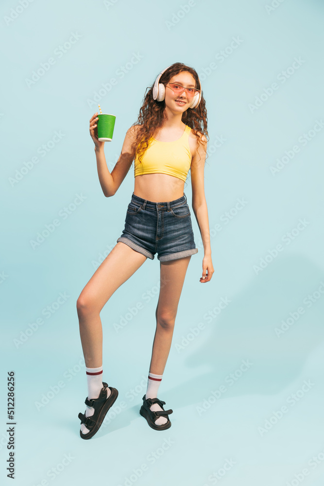 Portrait of young girl wearing shorts, yellow top and headphones, posing with coffee cup isolated over light blue background Portrait of young girl wearing shorts, yellow top and headphones, posing with coffee cup isolated over light blue background