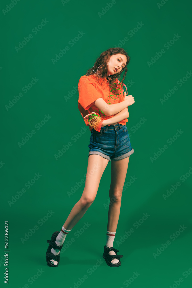 Portrait of tender young girl in casual outfit, shorts and colorful T-shirt posing isolated over green studio background Portrait of tender young girl in casual outfit, shorts and colorful T-shirt posing isolated over green studio background