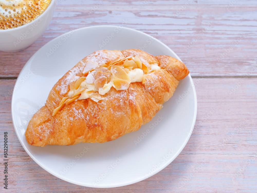 Croissant with cream and almonds on white circle plate and coffee cup over wooden table. Close up, space for text.. Croissant with cream and almonds on white circle plate and coffee cup over wooden table. Close up, space for text..