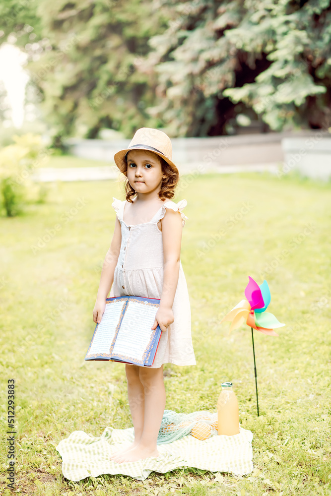 Cute little girl in a hat standing on a blanket on a park meadow and holding a book in a hands. Picnic in nature Cute little girl in a hat standing on a blanket on a park meadow and holding a book in a hands. Picnic in nature