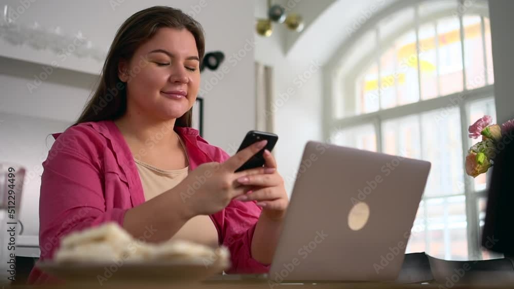 Young overweight woman is using phone and sitting at table with laptop in home office spbd. Close view of oversized European female holds smartphone and looks at display, types message and sits at Young overweight woman is using phone and sitting at table with laptop in home office spbd. Close view of oversized European female holds smartphone and looks at display, types message and sits at