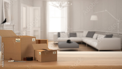 Wooden table, desk or shelf with stack of cardboard boxes over blurred view of classic living room with large sofa, modern interior design, moving house concept with copy space © ArchiVIZ