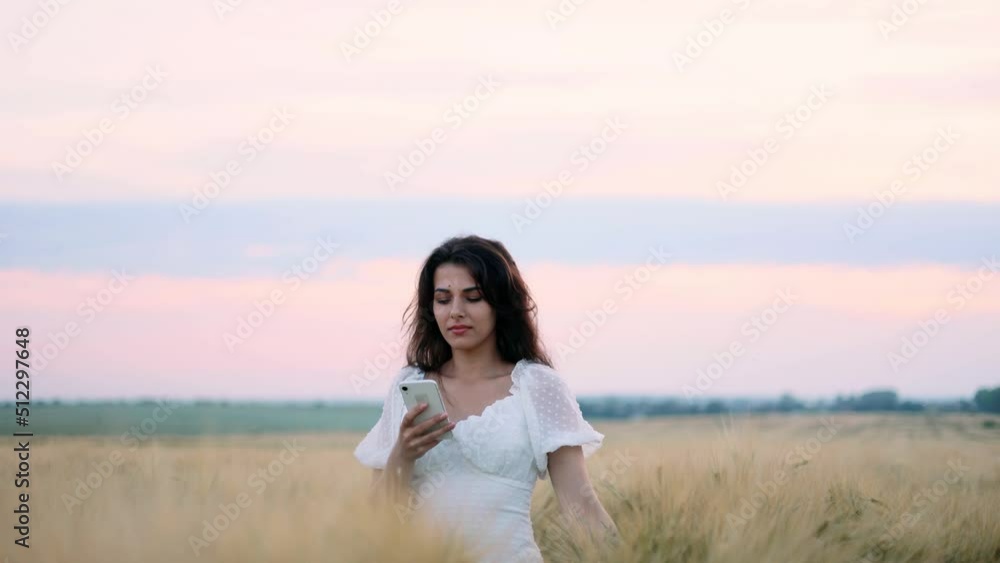 Young beatiful brunette woman in white dress texting on her phone and walking down in the gold field during the sunset. Young beatiful brunette woman in white dress texting on her phone and walking down in the gold field during the sunset.