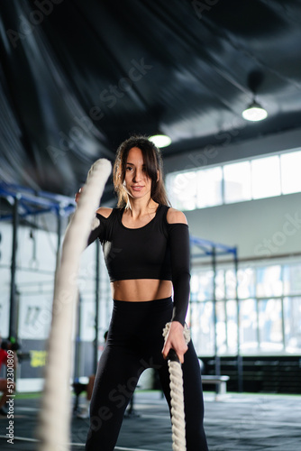 Powerful woman training battle ropes at cardio workout in dark gym. Professional athlete exercise fitness sport club equipment. Strong bodybuilder lifting weights. Athletic person effort