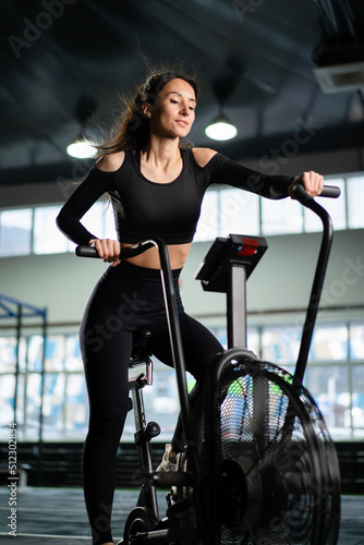 Young Athletic middle aged woman training on air resistance bike, cross training workout set in gym. Active woman spinning a air bike in gym with trainers. female training on air bike.