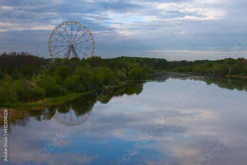 A high Ferris wheel stands on the bank of the river, in the water of which the evening sky is reflected