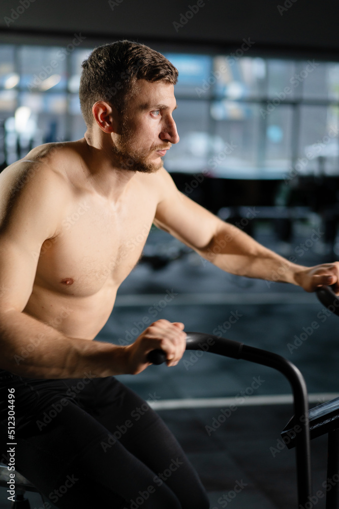 Obraz premium Young Athletic middle aged man training on air resistance bike, cross training workout set in gym. Active man spinning a air bike in gym with trainers. Male training on air bike.