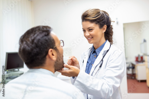 A young man sits on an exam table across from his doctor. The doctor reaches forward with a tongue depressor as the man looks up and sticks out his tongue.