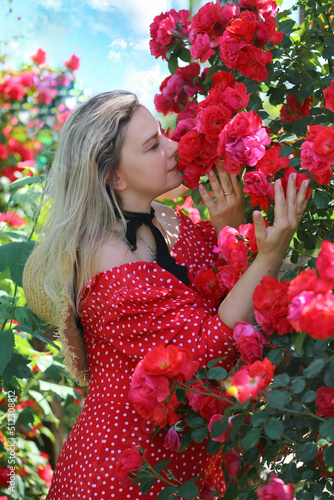 Cute Caucasian girl in a red dress with polka dots enjoying the scent of roses. Stunning flower garden backdrop. Flowers. Summer. Travel concept.
