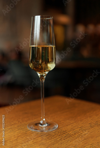 Glass of champagne standing on wooden table in soft focus on naturally blurred dark background. White wine with bubbles. Celebration, happy holidays concept