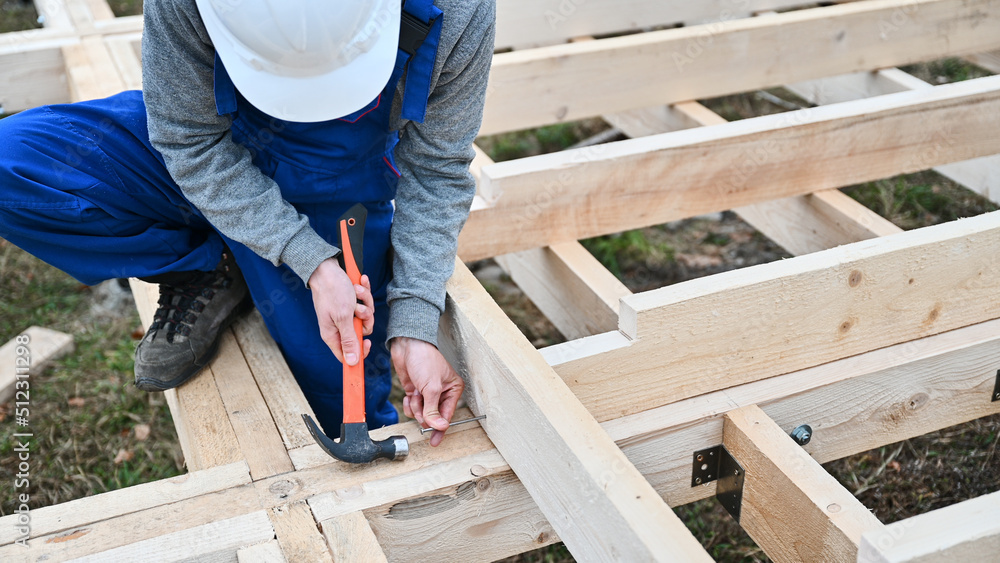 Man worker building wooden frame house on pile foundation. Carpenter hammering nail into wooden board, using hammer. Carpentry concept. Man worker building wooden frame house on pile foundation. Carpenter hammering nail into wooden board, using hammer. Carpentry concept.