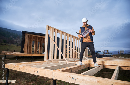 Male carpenter building wooden frame house on pile foundation. Man builder standing on construction site in safety helmet, with construction documentation inspecting quality of work.