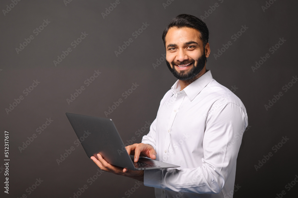 Indian male entrepreneur carrying laptop isolated on black, bearded businessman using laptop, typing, receiving emails, using new computer app, looking at the camera and smiles Indian male entrepreneur carrying laptop isolated on black, bearded businessman using laptop, typing, receiving emails, using new computer app, looking at the camera and smiles