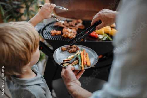 Unrecognizable father with little son grilling ribs and vegetable on grill during family summer garden party, close-up