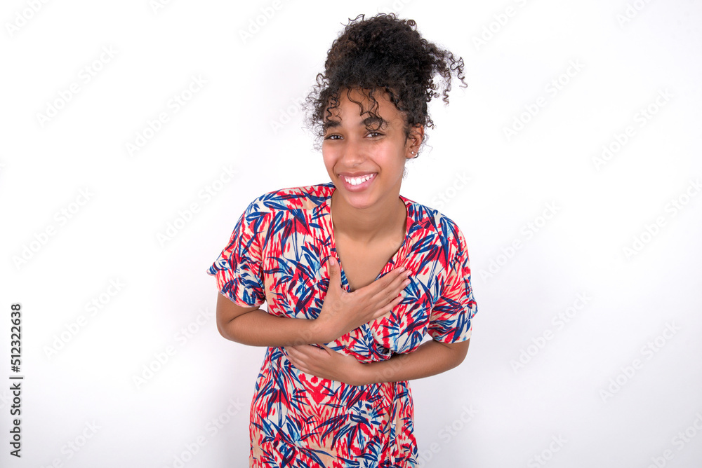 Young African American woman wearing colourful dress over white wall keeps hands crossed, laughs out loud at good joke, wears casual clothes. Young African American woman wearing colourful dress over white wall keeps hands crossed, laughs out loud at good joke, wears casual clothes.